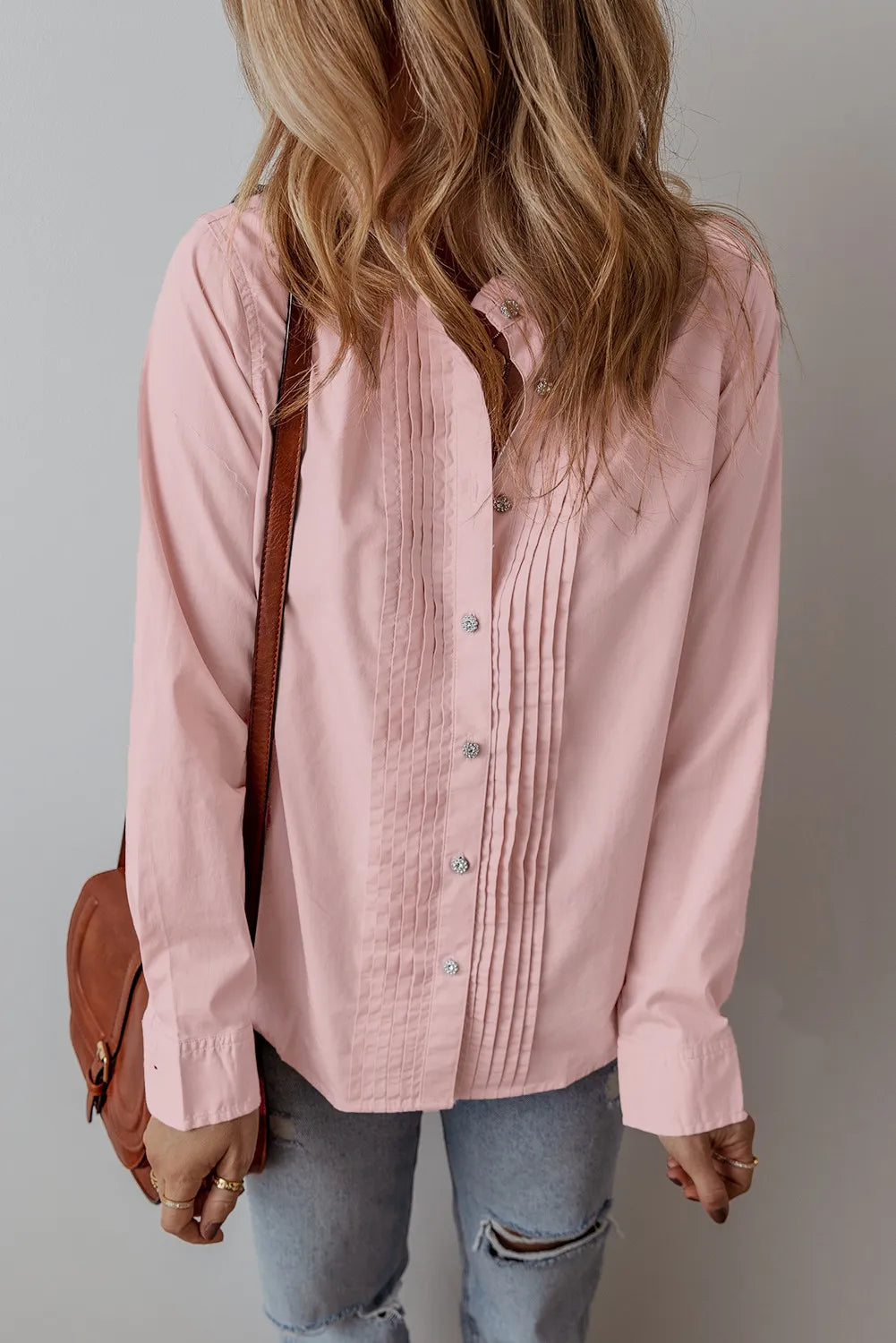 A person with long wavy hair is wearing the Long Sleeve Shirt in blush pink, crafted from 100% cotton and showcasing pleated details, paired with light blue distressed jeans. They have a brown leather bag slung over their right shoulder against a plain light gray wall. For best care, machine wash cold and tumble dry low.