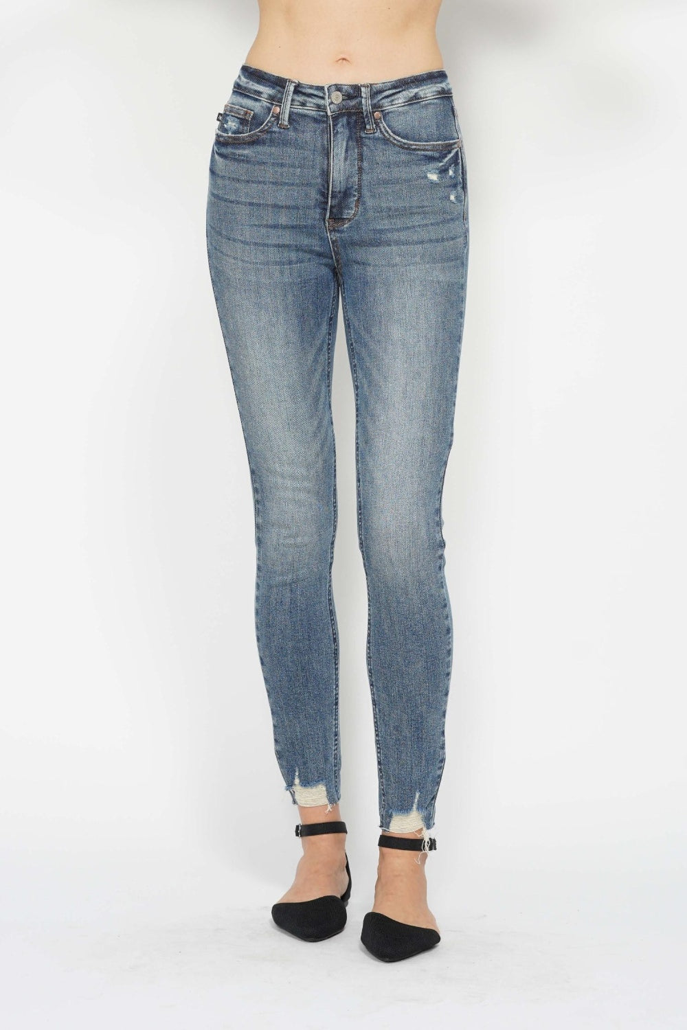 A person is wearing the Judy Blue Tummy Control Vintage Wash Hem Destroy Skinny Jeans, which are high-waisted with slight distressing on the front pockets and feature frayed hems at the ankles. The jeans are styled with black ankle-strap flats, and the plain white background highlights their fit and style.