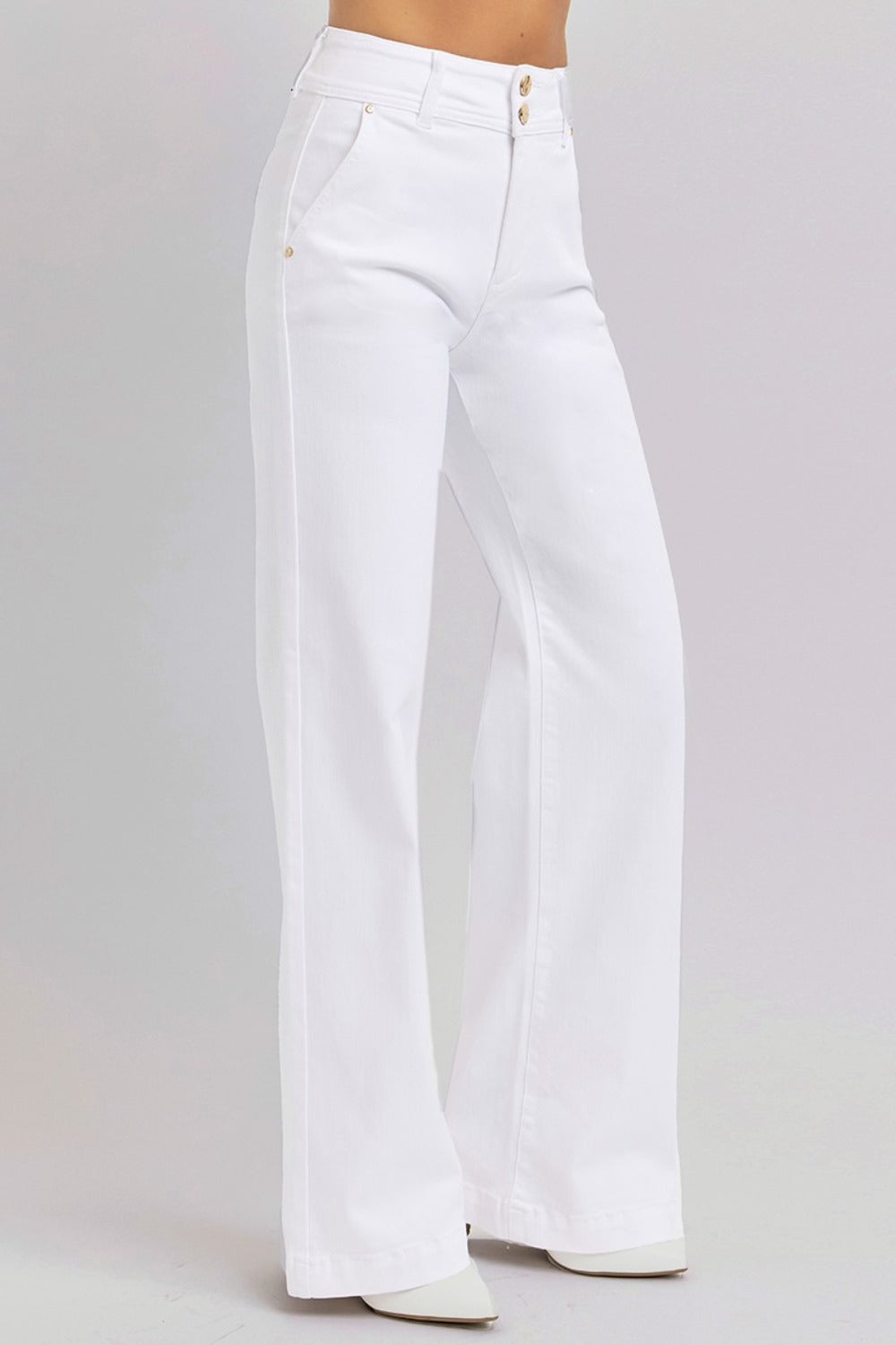 A person stands on a light background wearing the RISEN Tummy Control Double Button Wide Leg Jeans, characterized by their high-waisted, wide-leg design in white. These jeans feature side pockets and are paired with white pointed-toe shoes, delivering a smooth, seamless appearance that complements the clean and minimalistic style.