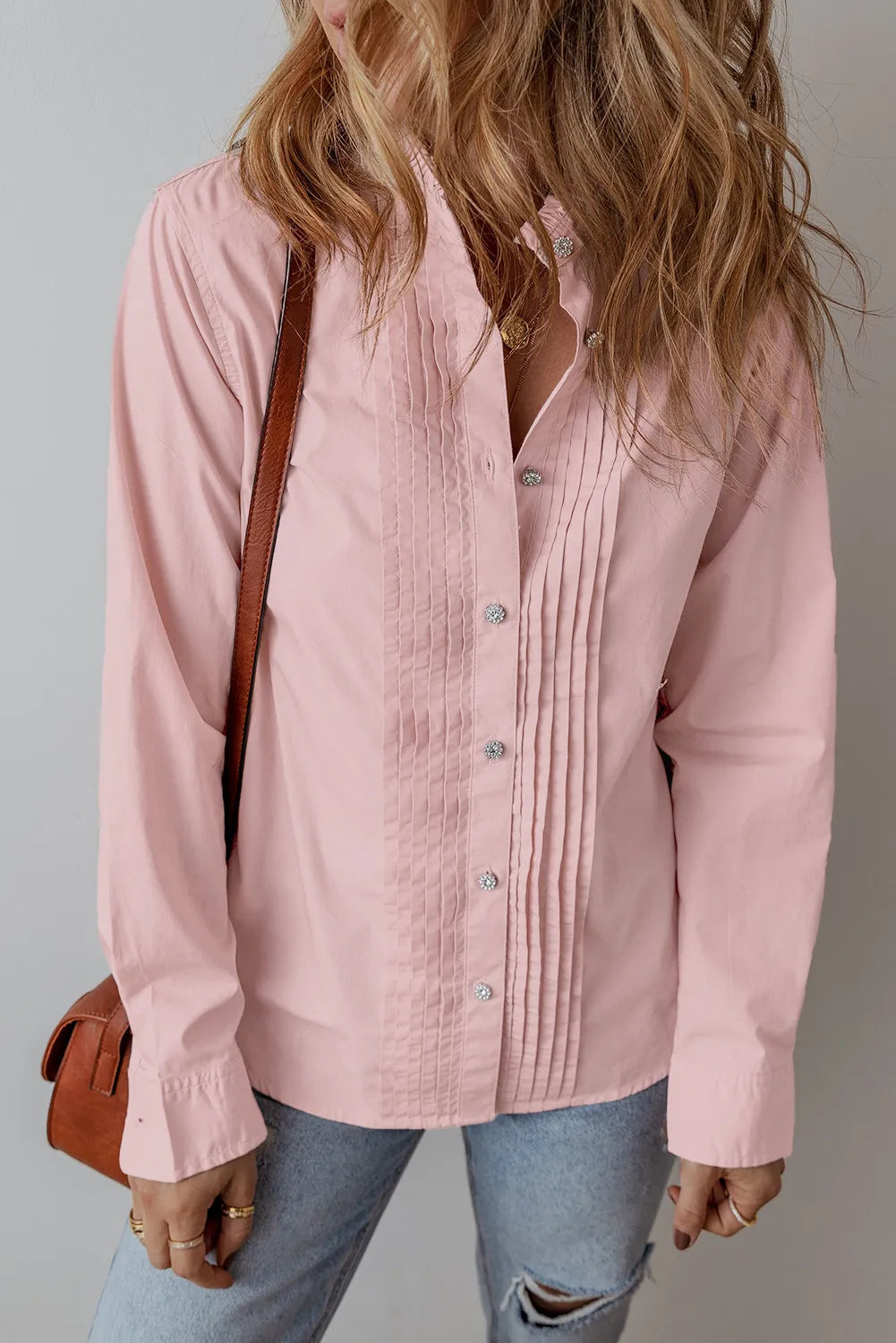 A woman with wavy hair is dressed in a blush pink, 100% cotton button-up shirt known as the Long Sleeve Shirt, which features vertical pleats along the front. She pairs it with light distressed jeans and carries a brown leather shoulder bag. The photo is cropped at the neck and waist, set against a simple light gray background.