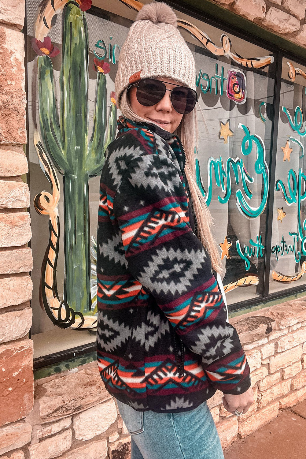 Woman wearing featured Aztec Printed Zip Up Jacket in Black and grey, paired with jeans, beige beanie, and sunglasses. Side view