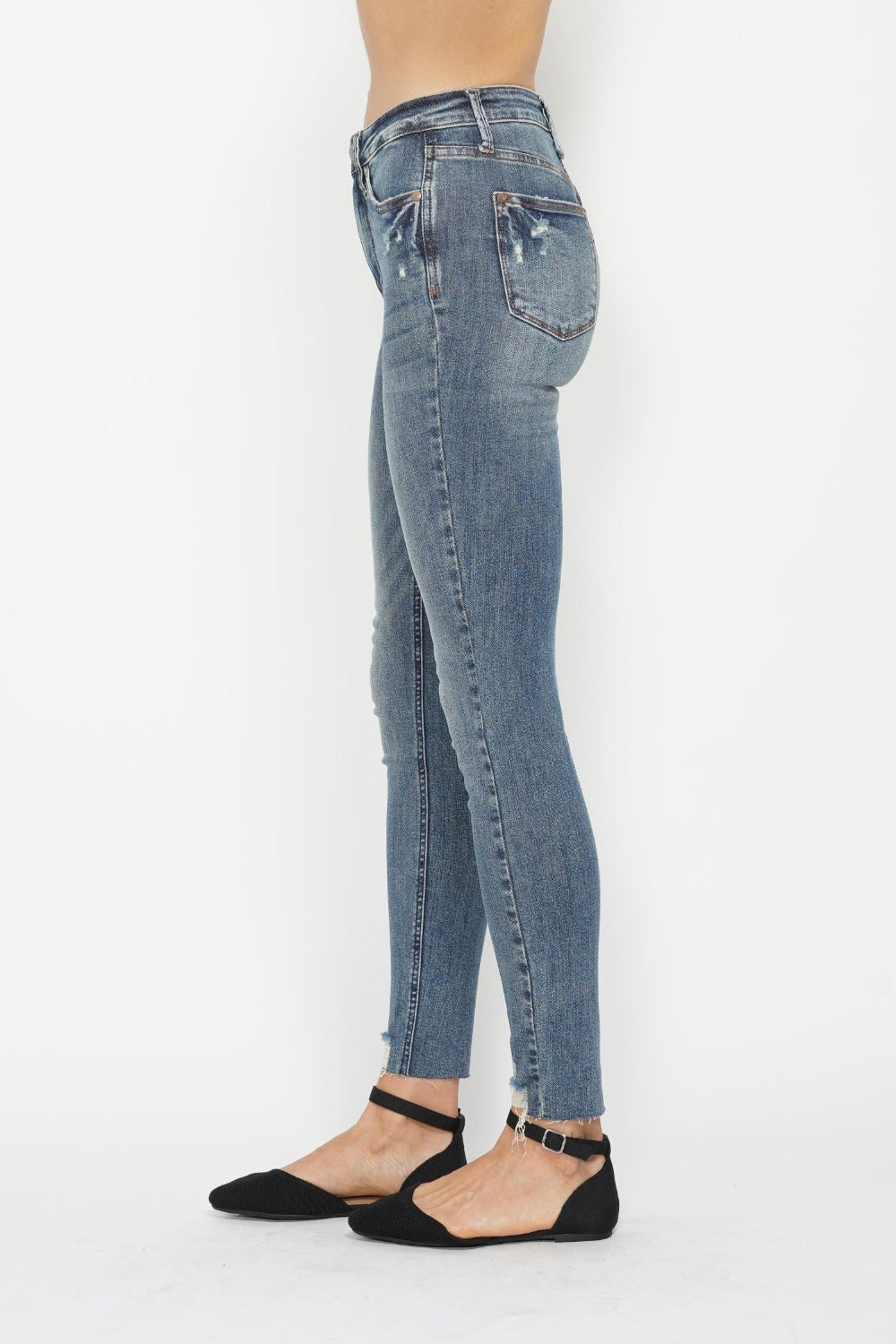 A side profile showcases a person wearing Judy Blue Tummy Control Vintage Wash Hem Destroy Skinny Jeans, characterized by their high waist, distressed look, and frayed hem. These jeans offer a slightly worn appearance with tummy control and are complemented by black pointed-toe flat shoes featuring ankle straps. The outfit is set against a plain white background.