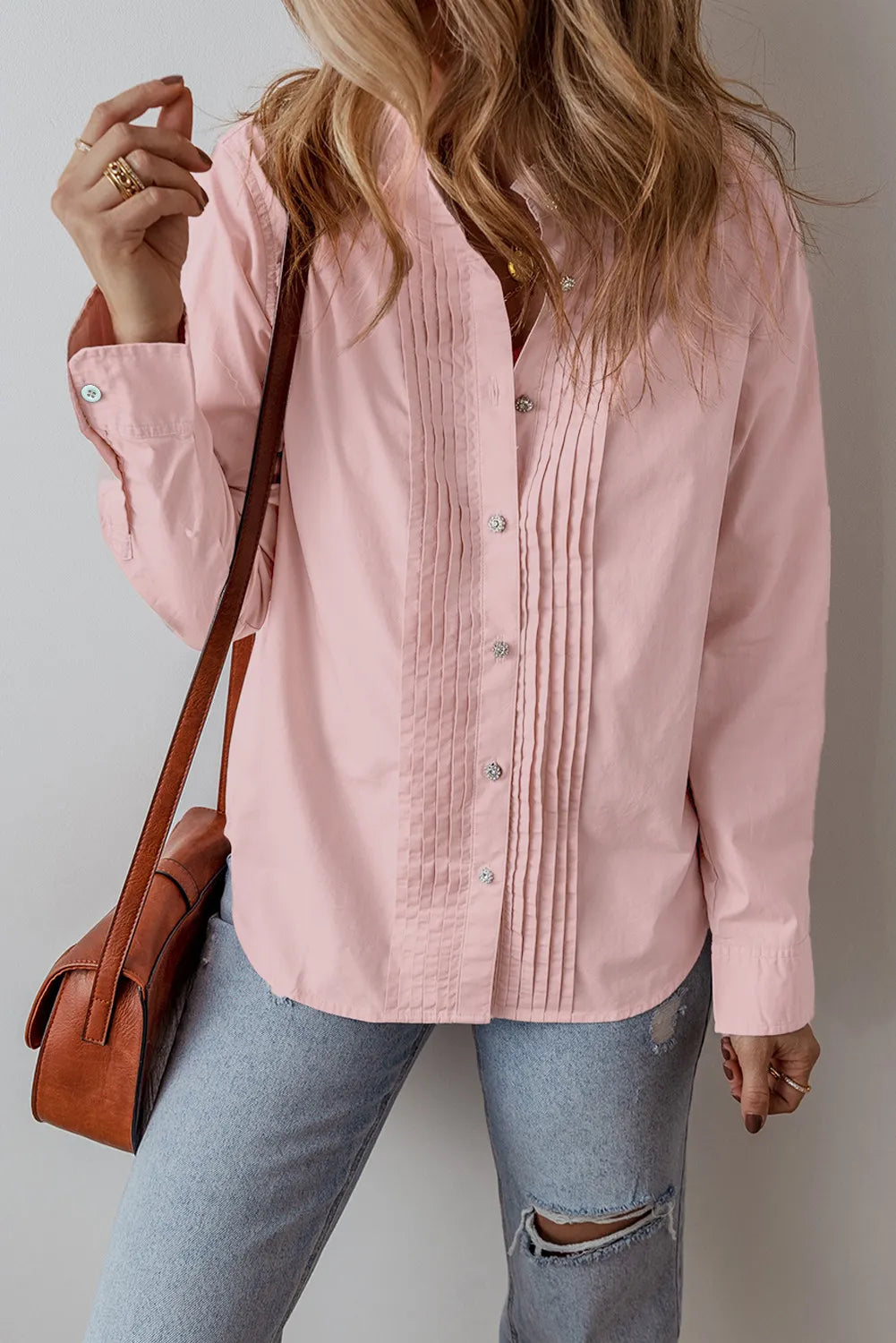 A woman with wavy blonde hair wears a blush pink long sleeve shirt made of 100% cotton and distressed light blue jeans. She carries a brown leather shoulder bag, adorned with rings on her fingers. Her hand is raised near her face as she stands against a neutral background.