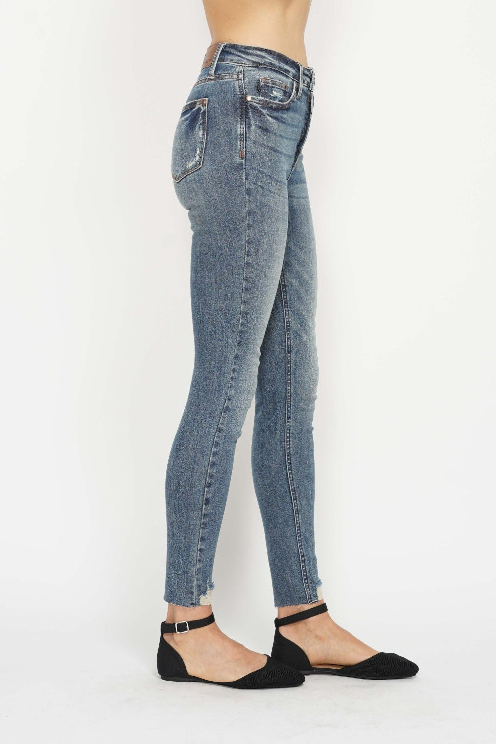 A person is shown from the side against a white background, wearing Judy Blue Tummy Control Vintage Wash Hem Destroy Skinny Jeans, which are high-waisted with a distressed look and frayed hems. They are paired with black pointed-toe flats that have ankle straps.