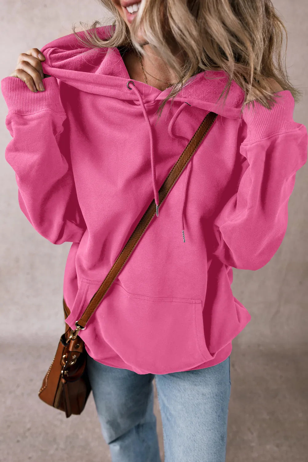 A person in a bright pink drawstring dropped shoulder hoodie with the hood partially up, paired with light blue jeans. They carry a brown crossbody bag. The neutral-toned background contrasts their smile, visible only on the lower half of their face.