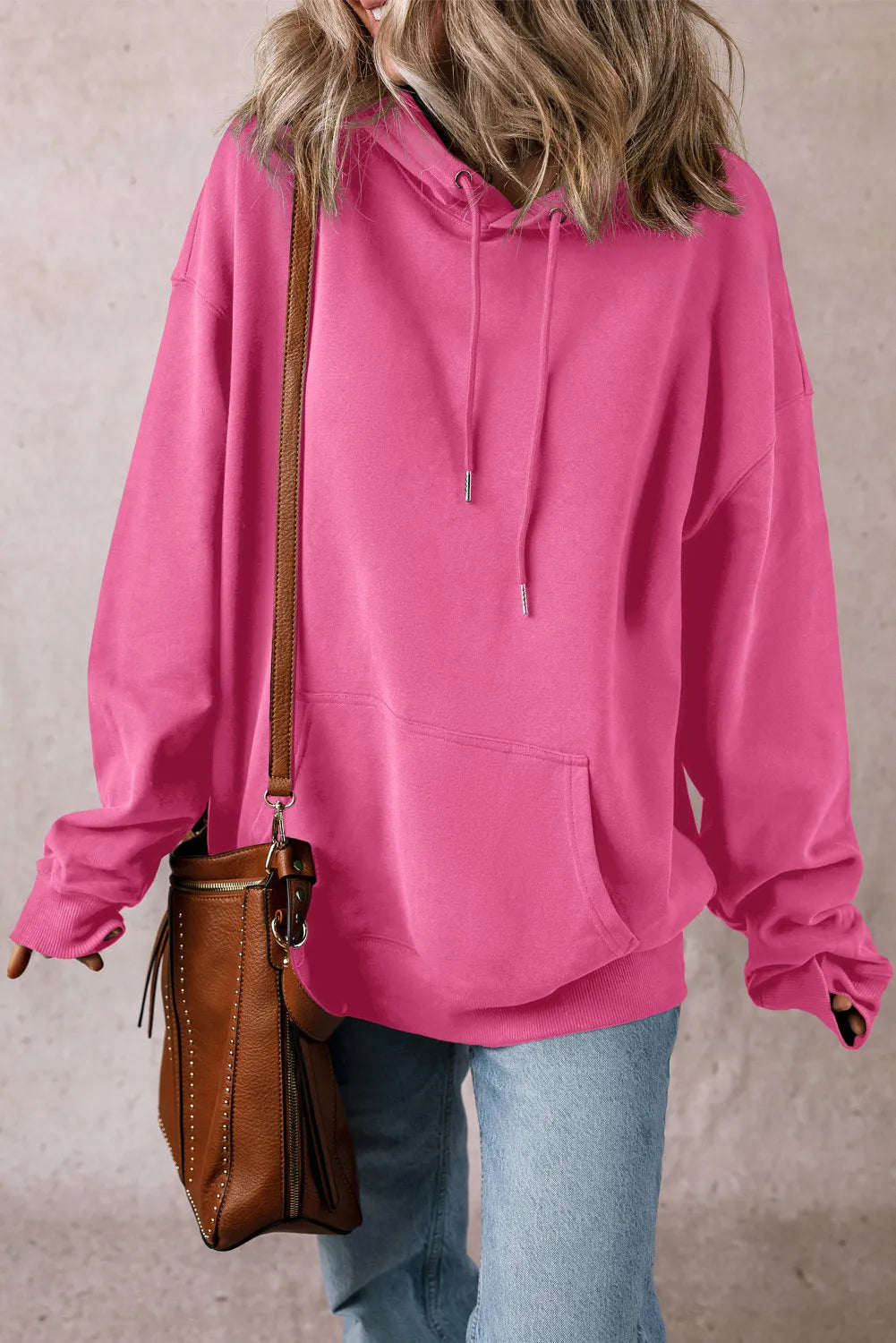 A person wearing a loose-fitting, pink "Drawstring Dropped Shoulder Hoodie" made of polyester, along with light blue jeans, carries a brown leather shoulder bag adorned with fringe details. Their head is cropped out, and their hair slightly obscures the hood. The background features a simple, textured wall in a light shade.