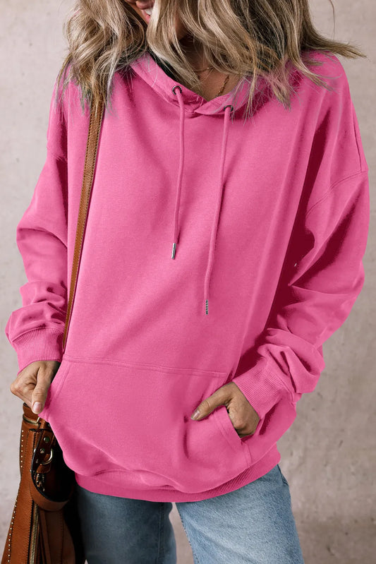 A person wearing a bright pink Drawstring Dropped Shoulder Hoodie with long sleeves and a front pocket complements their light blue jeans. They carry a brown leather shoulder bag against the plain, light-colored background.