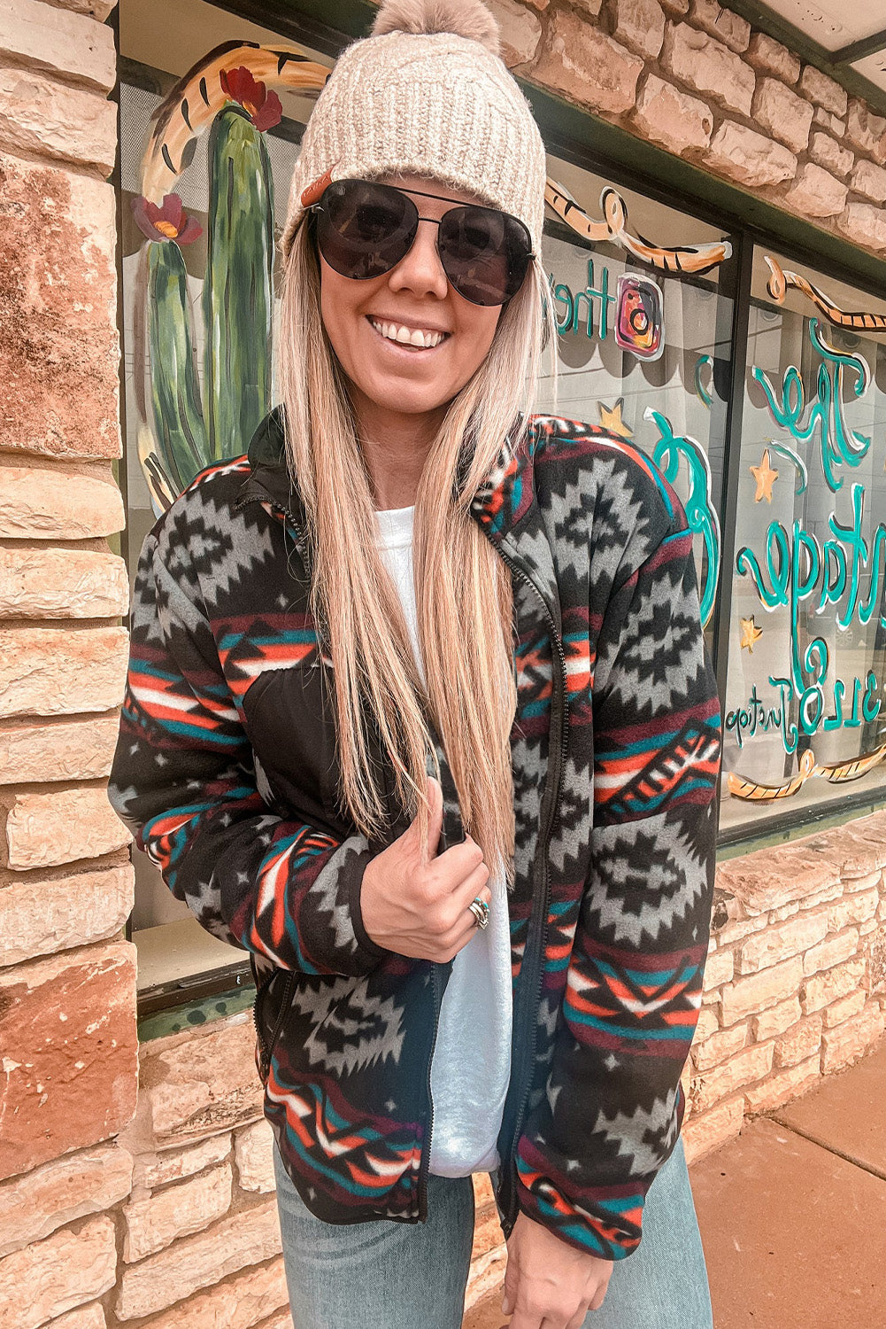 Woman wearing featured Aztec Printed Zip Up Jacket in Black and grey, paired with jeans, t-shirt, beige beanie, and sunglasses. front view.