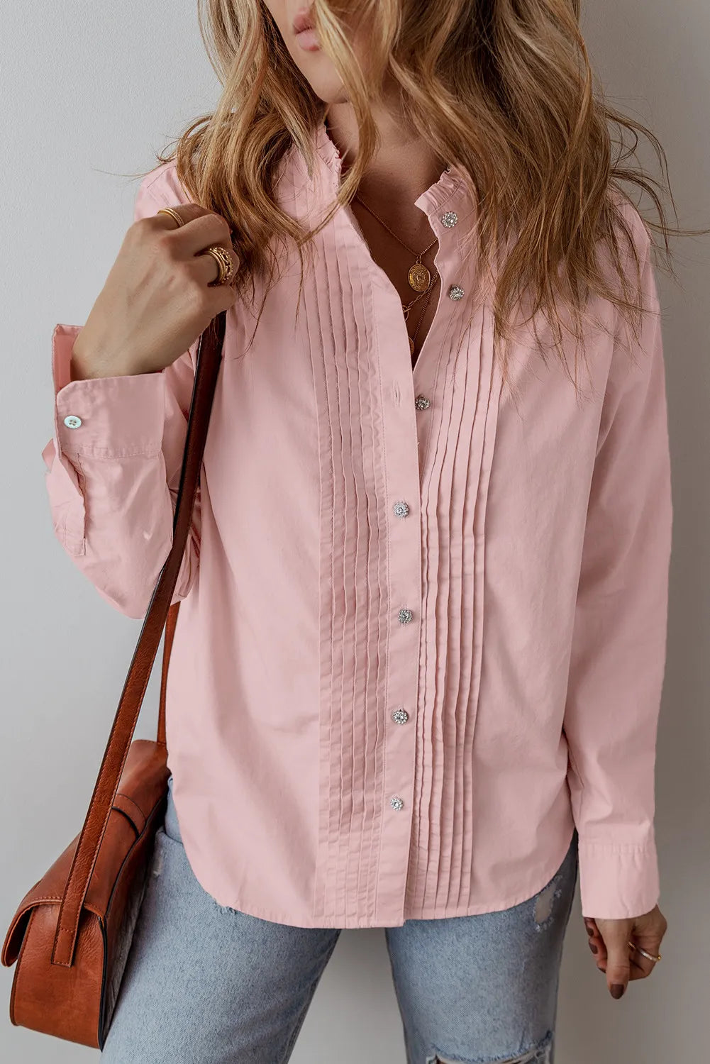 A woman stands near a white wall, wearing a Long Sleeve Shirt in Blush Pink made of 100% cotton, embellished with pleats beside the buttons. Her long, wavy light brown hair pairs beautifully with her faded blue jeans. She carries a brown leather crossbody bag and wears multiple rings with manicured nails.