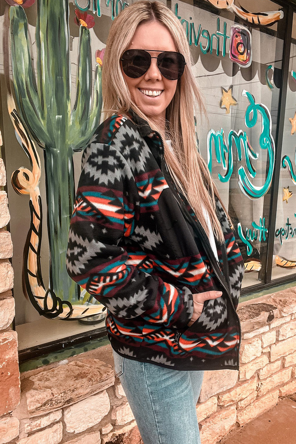 Woman wearing featured Aztec Printed Zip Up Jacket in Black and grey, paired with jeans and a t-shirt. Side view.