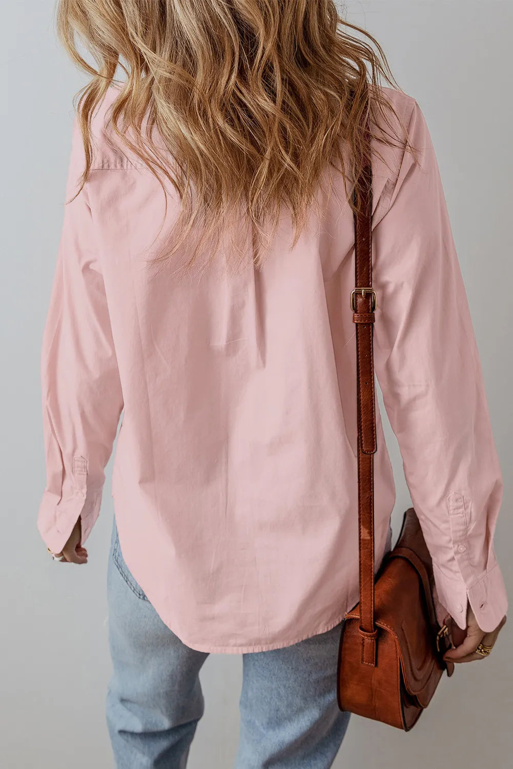 A person with wavy blonde hair, dressed in a Long Sleeve Shirt - Blush Pink made of 100% cotton and light blue jeans, stands facing away. They carry a brown leather shoulder bag over their left arm against a plain, light-colored background.