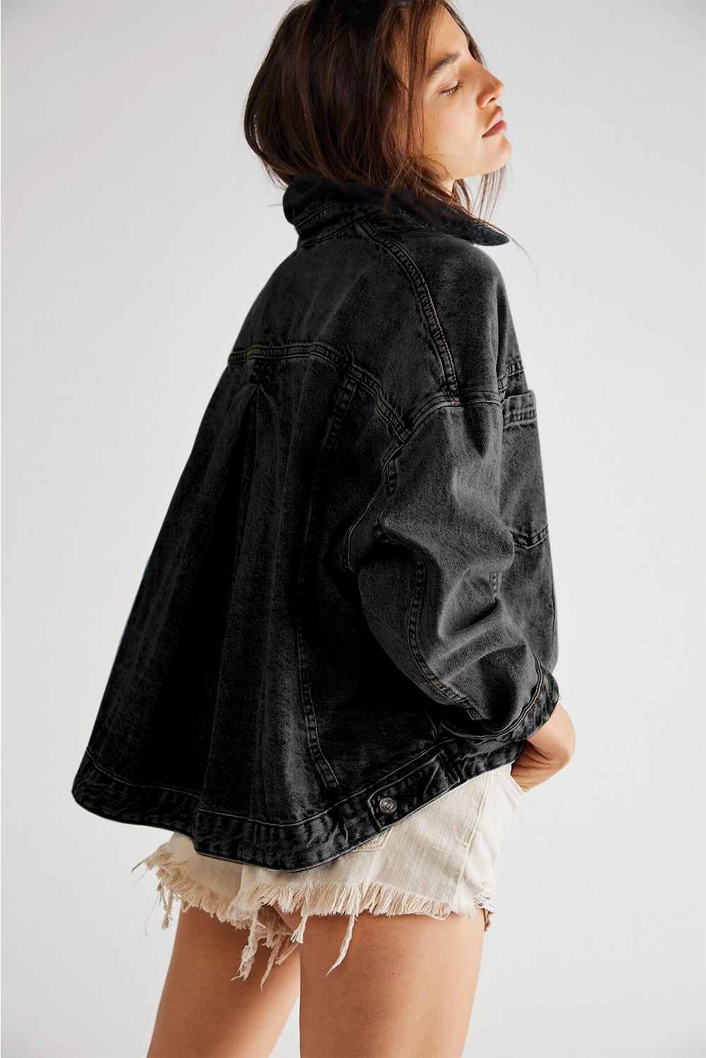 Dark-haired woman wearing the Black washed denim jacket. Oversized with button closure, turn down collar, and pockets. back left view