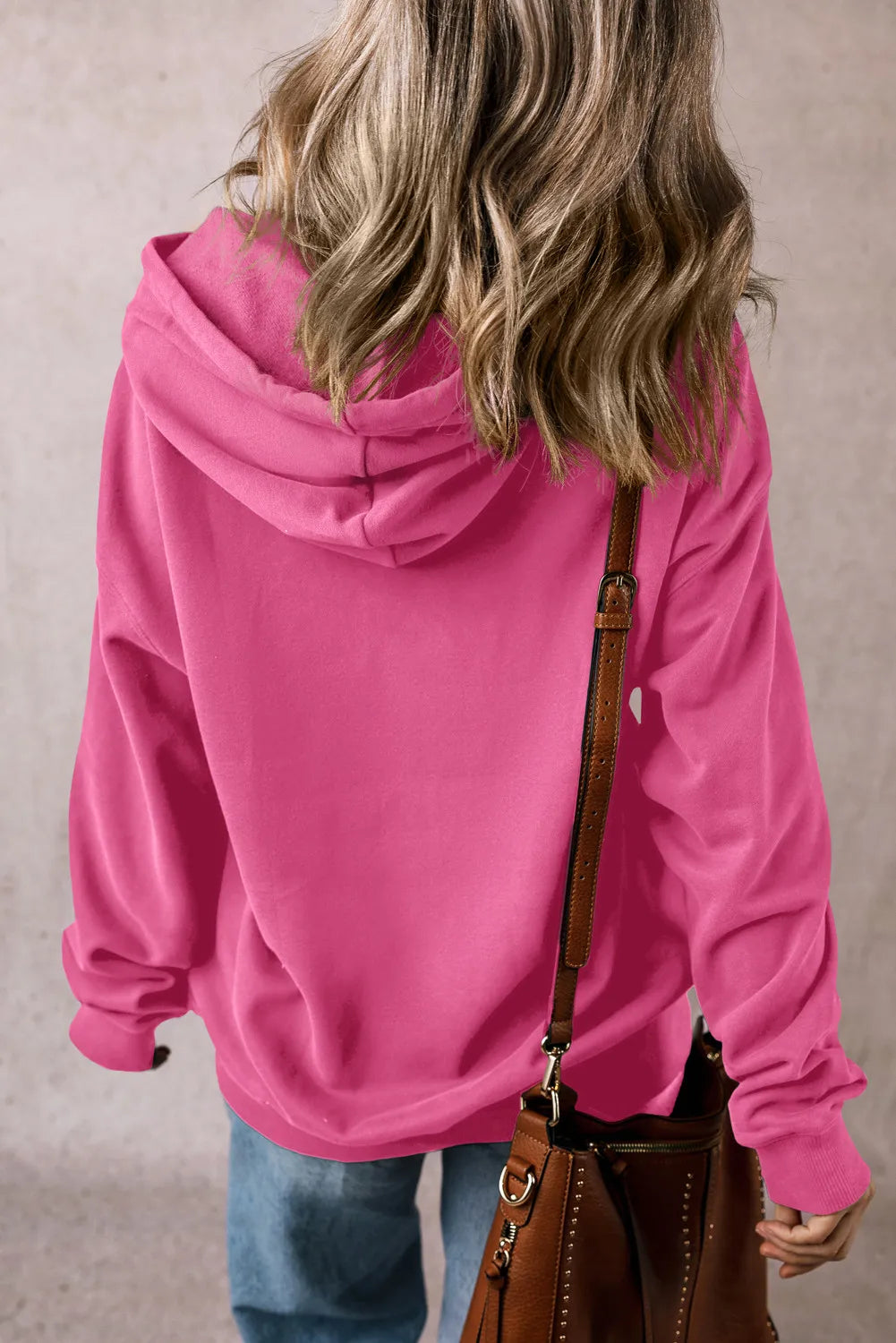 A person with wavy blonde hair is wearing a bright pink drawstring dropped shoulder hoodie and blue jeans, facing away. They carry a brown leather shoulder bag with metal accents. The background is a plain, textured surface.