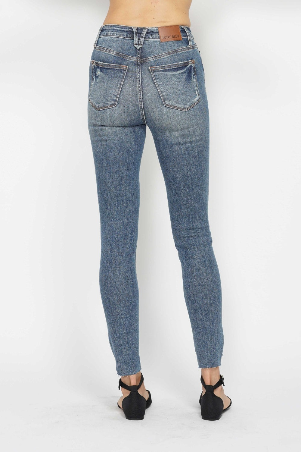 The image depicts someone from the back wearing Judy Blue Tummy Control Vintage Wash Hem Destroy Skinny Jeans, characterized by their high-waisted fit and vintage wash. The jeans have a label on the waistband, two back pockets with visible stitching, and a fitted cut. Additionally, the person is paired them with black sandals that feature low heels against a plain white background.